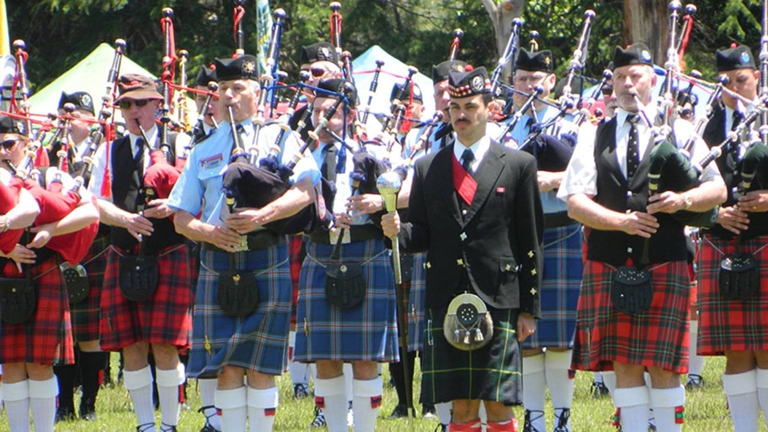 Band members blowing bagpipse while standing in formation at a park.