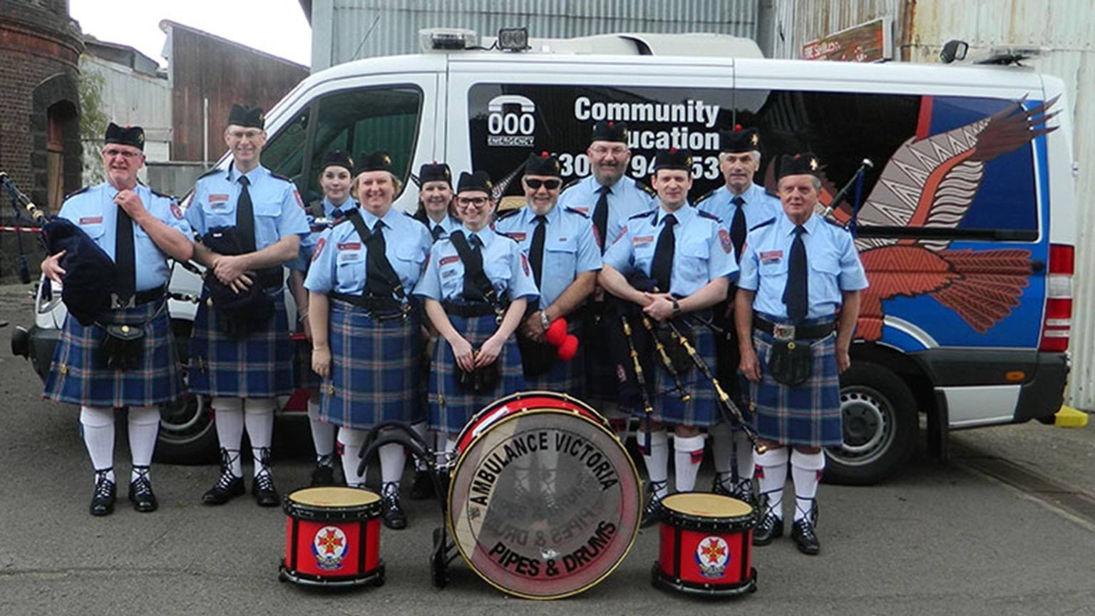 A group of band members standing beside an ambulance. Drum instruments are placed on the ground in front of them.