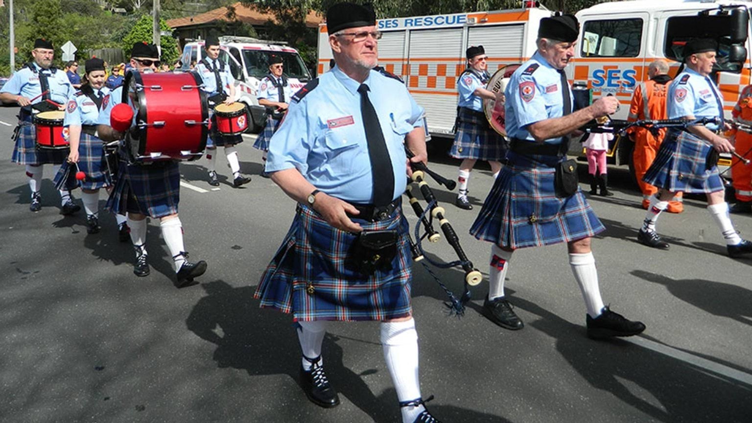 Band members playing the drum while marching down the street. Emergency response vehicles are parked alongside the road.