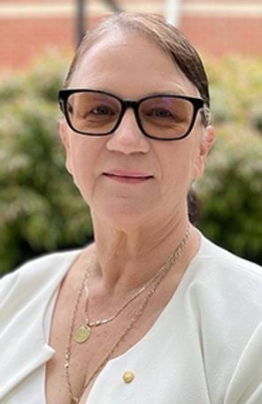 A woman with dark hair, wearing glasses and a white top, is standing outdoors in front of greenery.