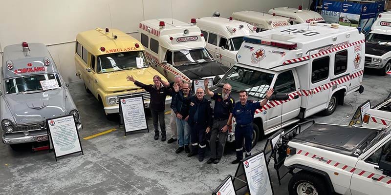 A group of men wave to the camera. They are surrounded by vintage ambulances.
