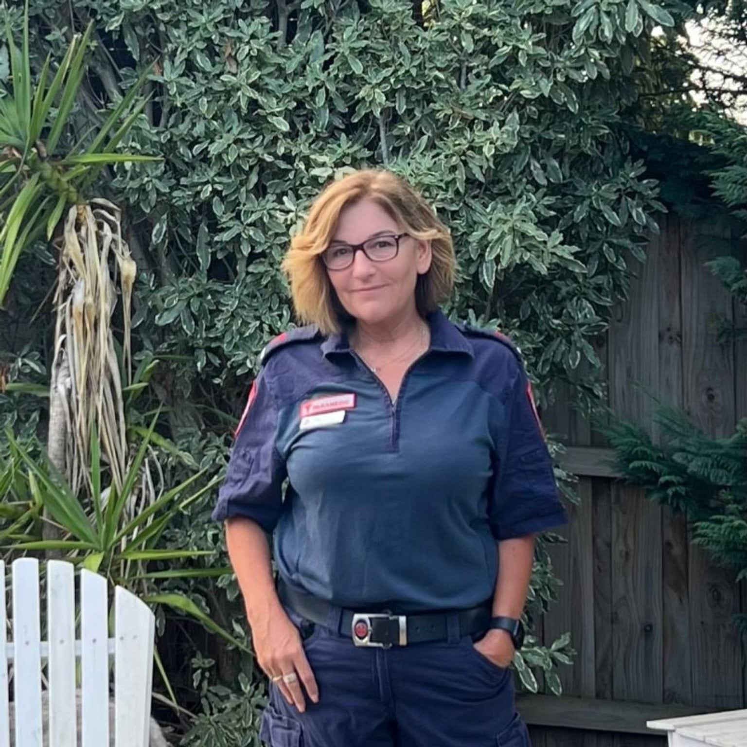 A woman in paramedic uniform smiles for the camera in front of a garden.