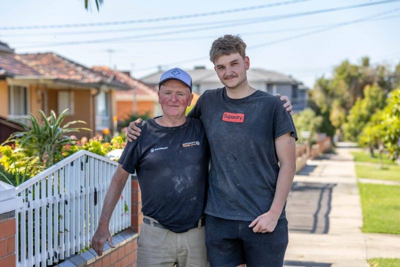 Two tradesmen smile for the camera with their arms around each other. They stand outside on the footpath of a residential street.