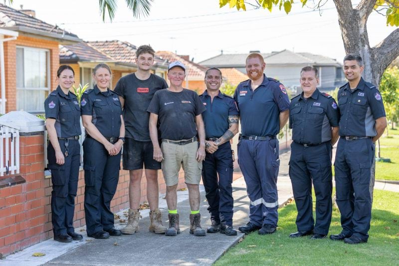 A group of firefighters and paramedics smile for the camera, with two tradesmen standing between them. They are on the footpath of a residential street.