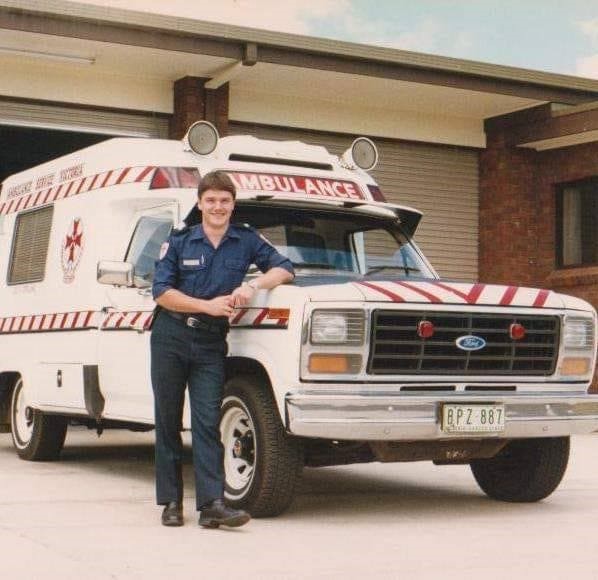 A man in ambulance uniform leans against an old-fashioned ambulance and smiles for the camera. It is a colour photo.