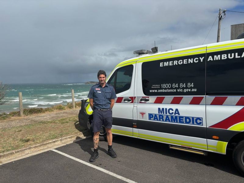 A man in paramedic uniform stands in front of a MICA vehicle. It's parked at a beach carpark. The ocean and grey sky are in the background.