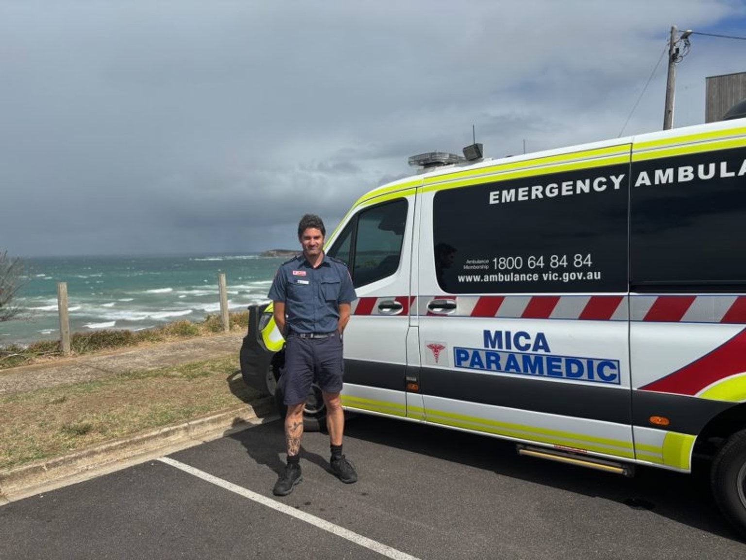 A man in paramedic uniform stands in front of a MICA vehicle. It's parked at a beach carpark. The ocean and grey sky are in the background.