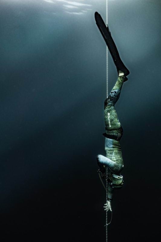 An underwater photo of a person in a full body wetsuit and flippers, swimming straight down.