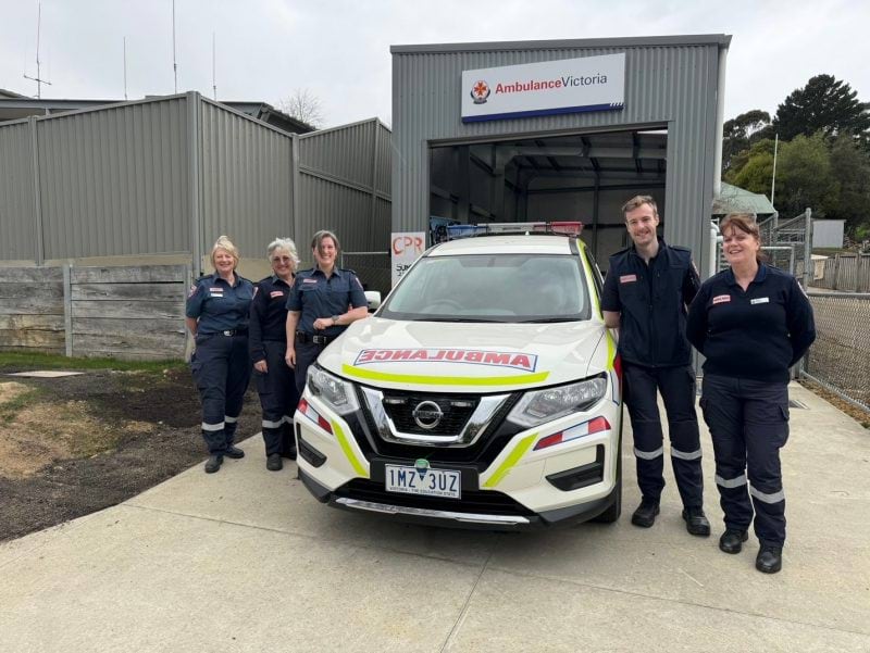 Five people in Ambulance Victoria uniform smile for the camera. They stand around an ambulance car in front of an Ambulance Victoria grey shed