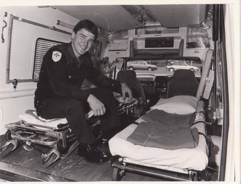 A black and white photo of a man in ambulance uniform, sitting inside an old-fashioned ambulance with two stretchers.