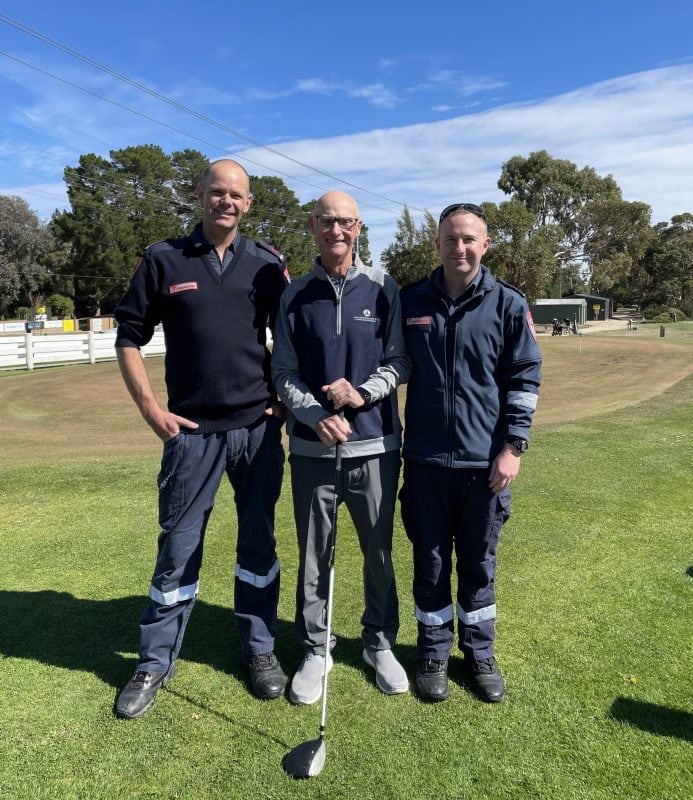 Two paramedics and a man in casual clothes smile for the camera in front of a golf green. The man in casual clothes holds a golf club.