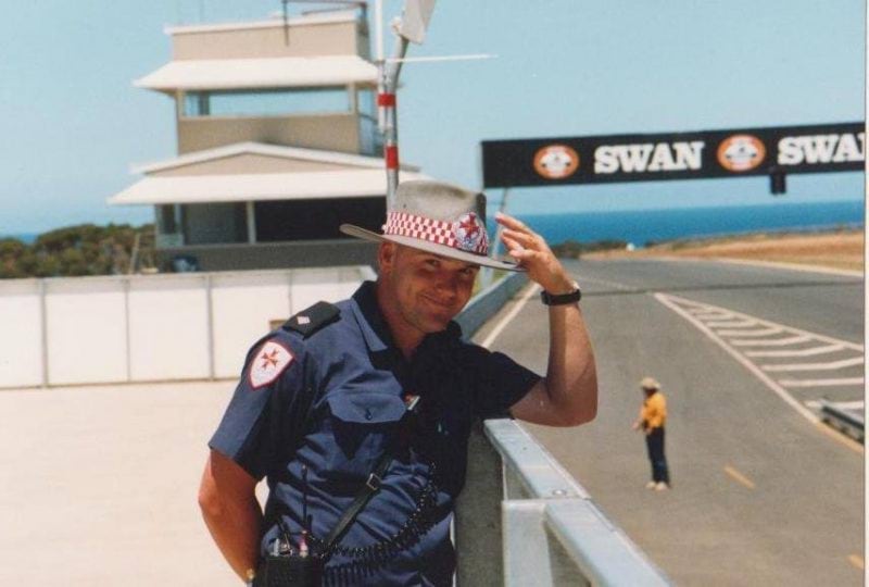 A man in ambulance uniform and ambulance broad brimmed hat smiles for the camera. He leans on the barrier at the edge of a race track.