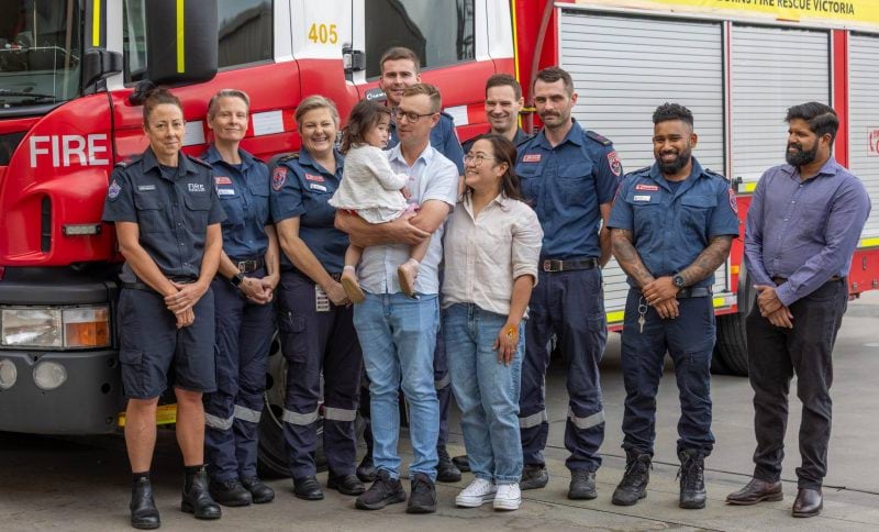 Paramedics, firefighters and a man in business wear smile for a photo with two parents and a little girl. They stand in front of a fire truck.