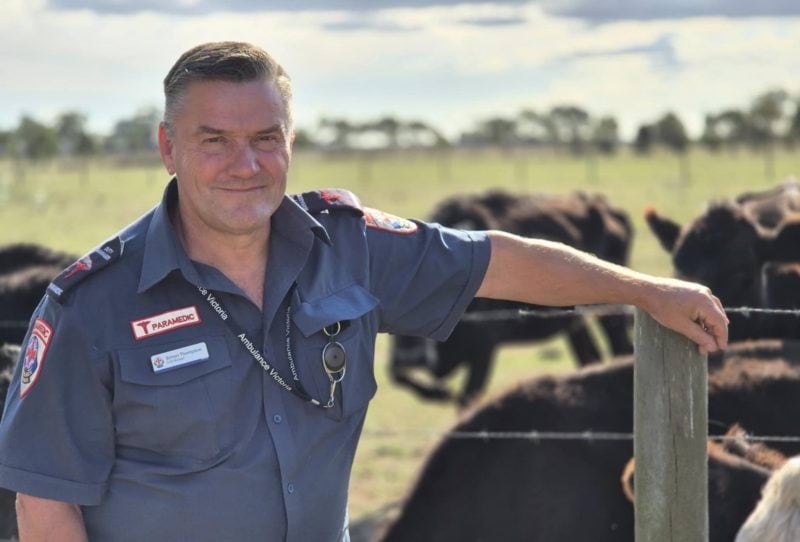 A male paramedic smiles for the camera. His arm rests on a farm fence, with black cows in a paddock behind him.