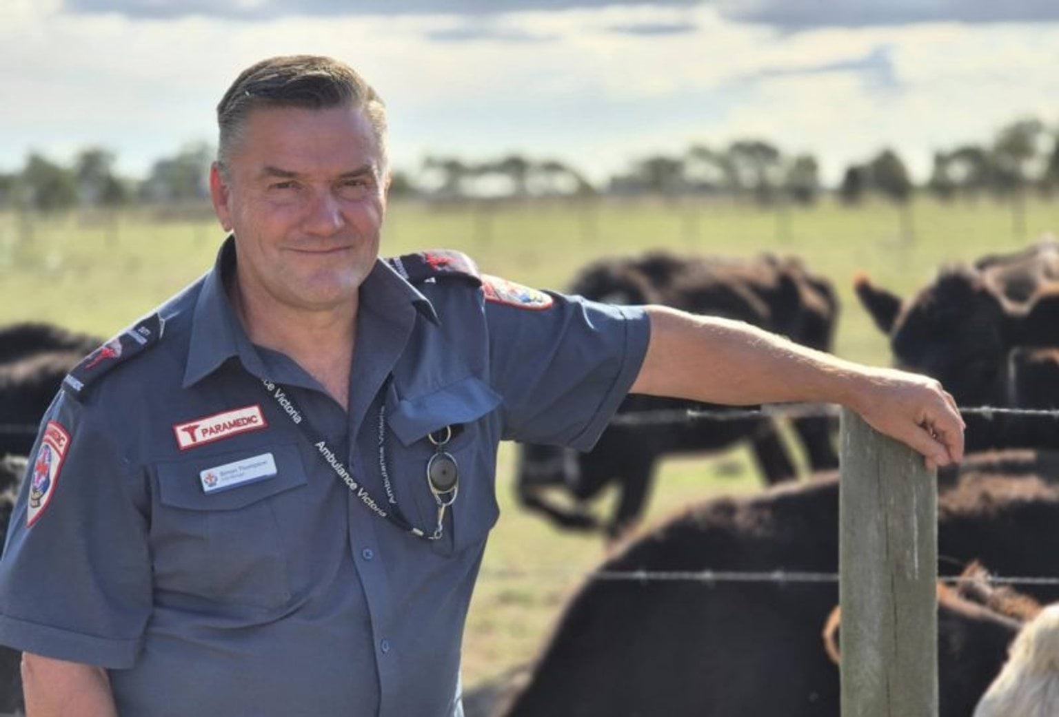 A male paramedic smiles for the camera. His arm rests on a farm fence, with black cows in a paddock behind him.