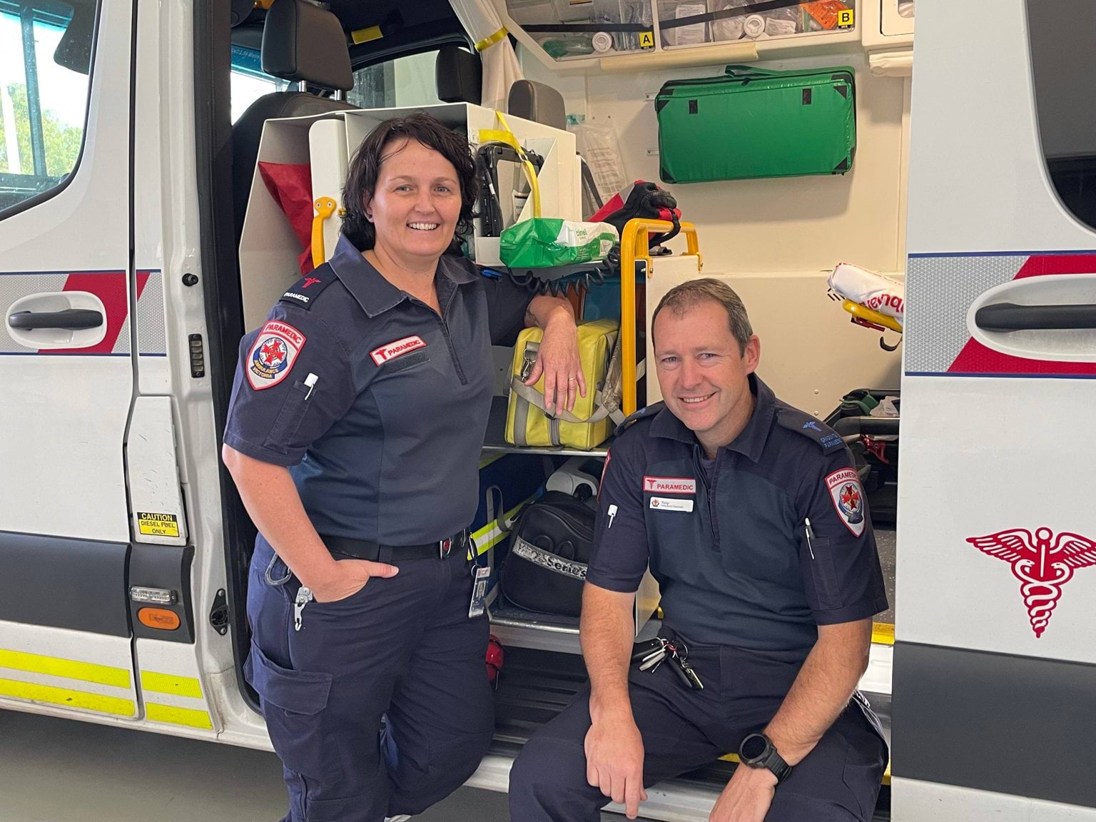 a woman and man smiling with an open ambulance behind them. 