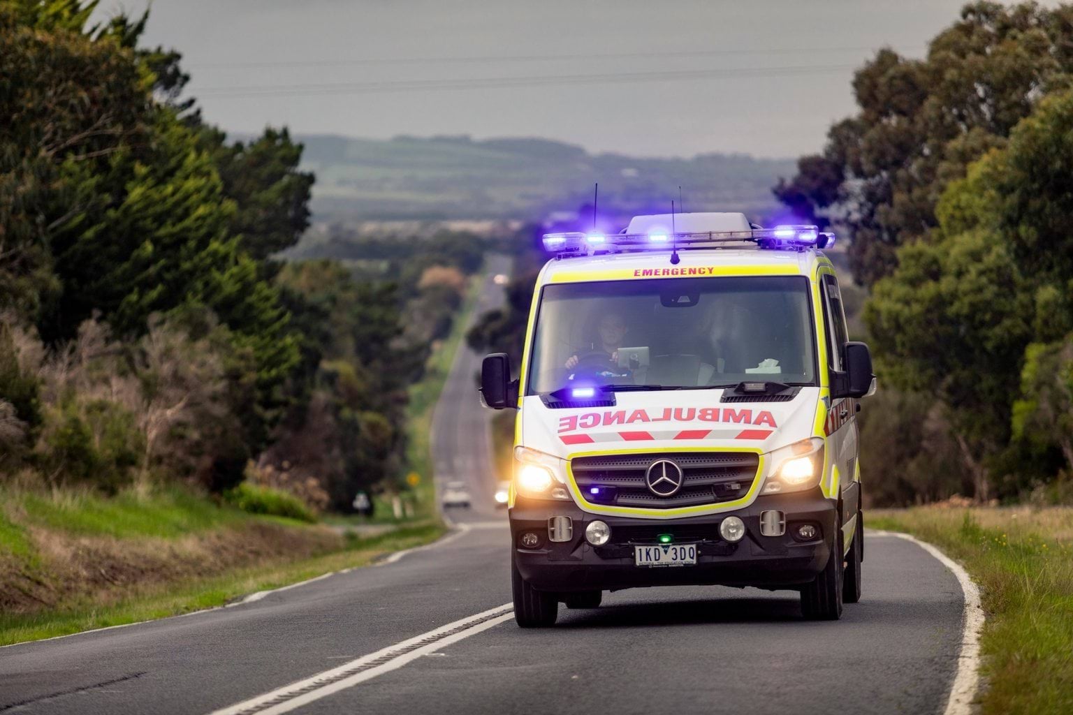 An ambulance with lights flashing drives up a hill on a long road.