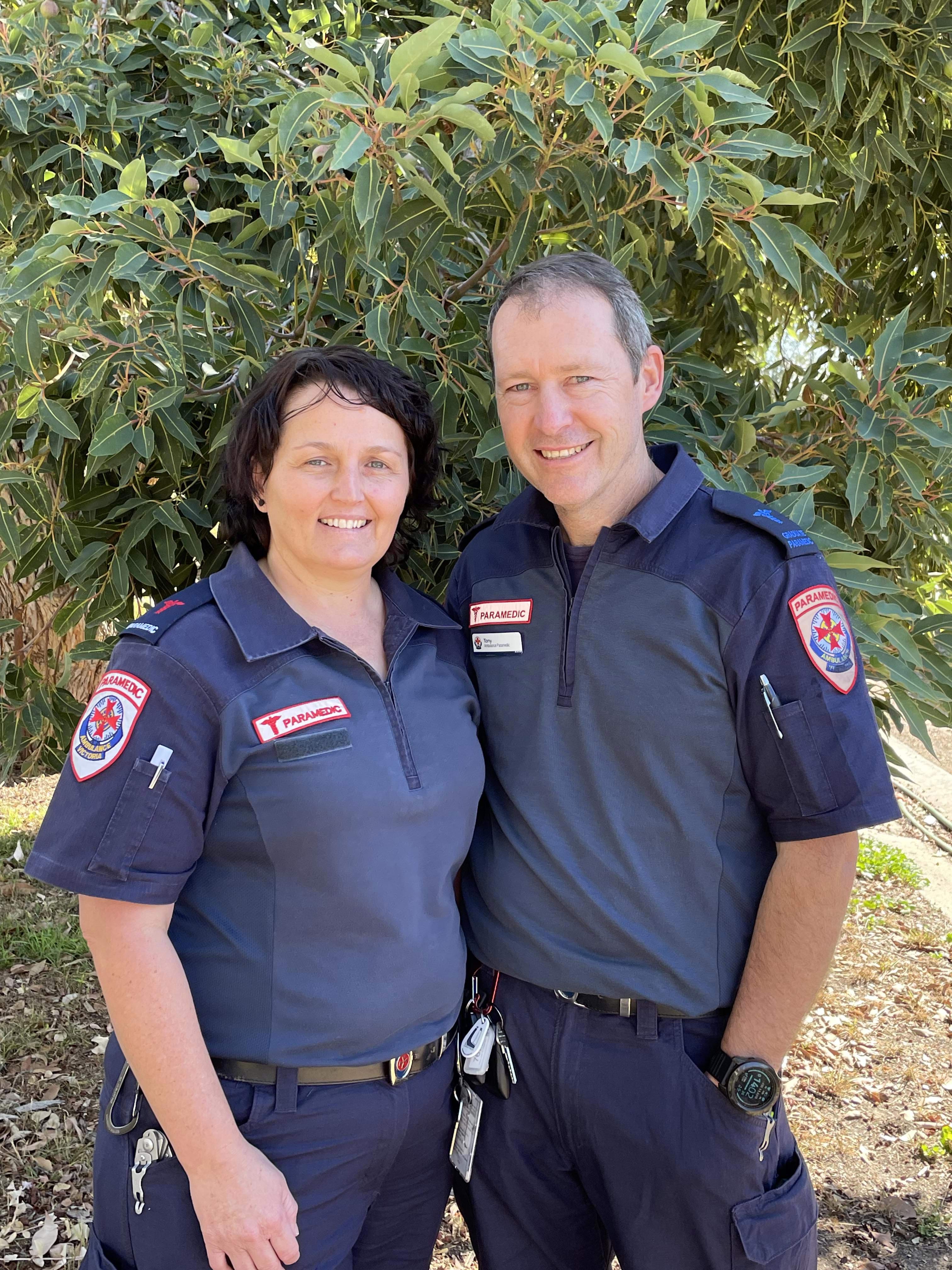 A woman and man in paramedic uniforms smiling in front of a tree.