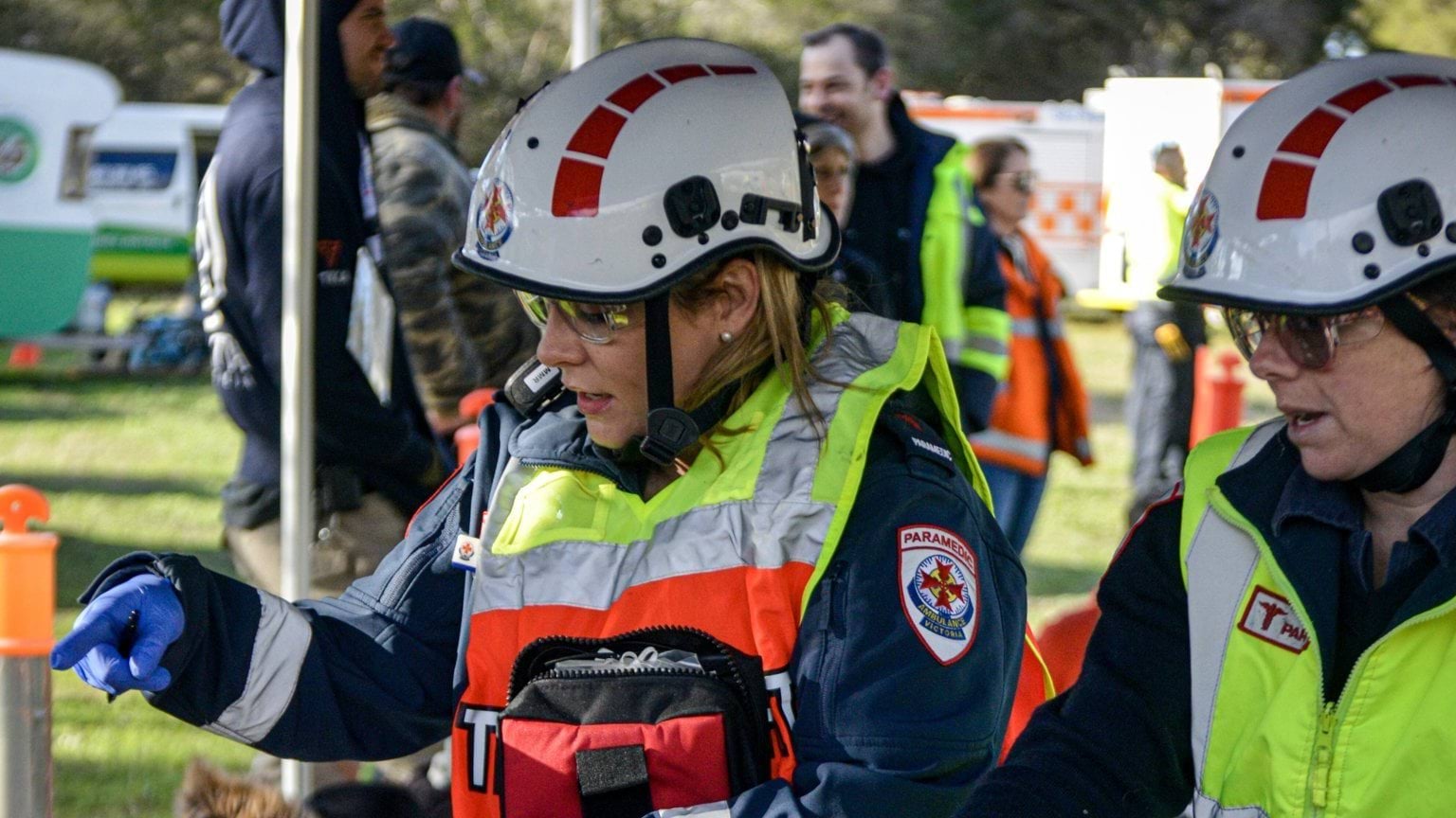 Two paramedics. There are people in casual clothes standing behind them. The paramedics are in discussion and one is pointing off camera.