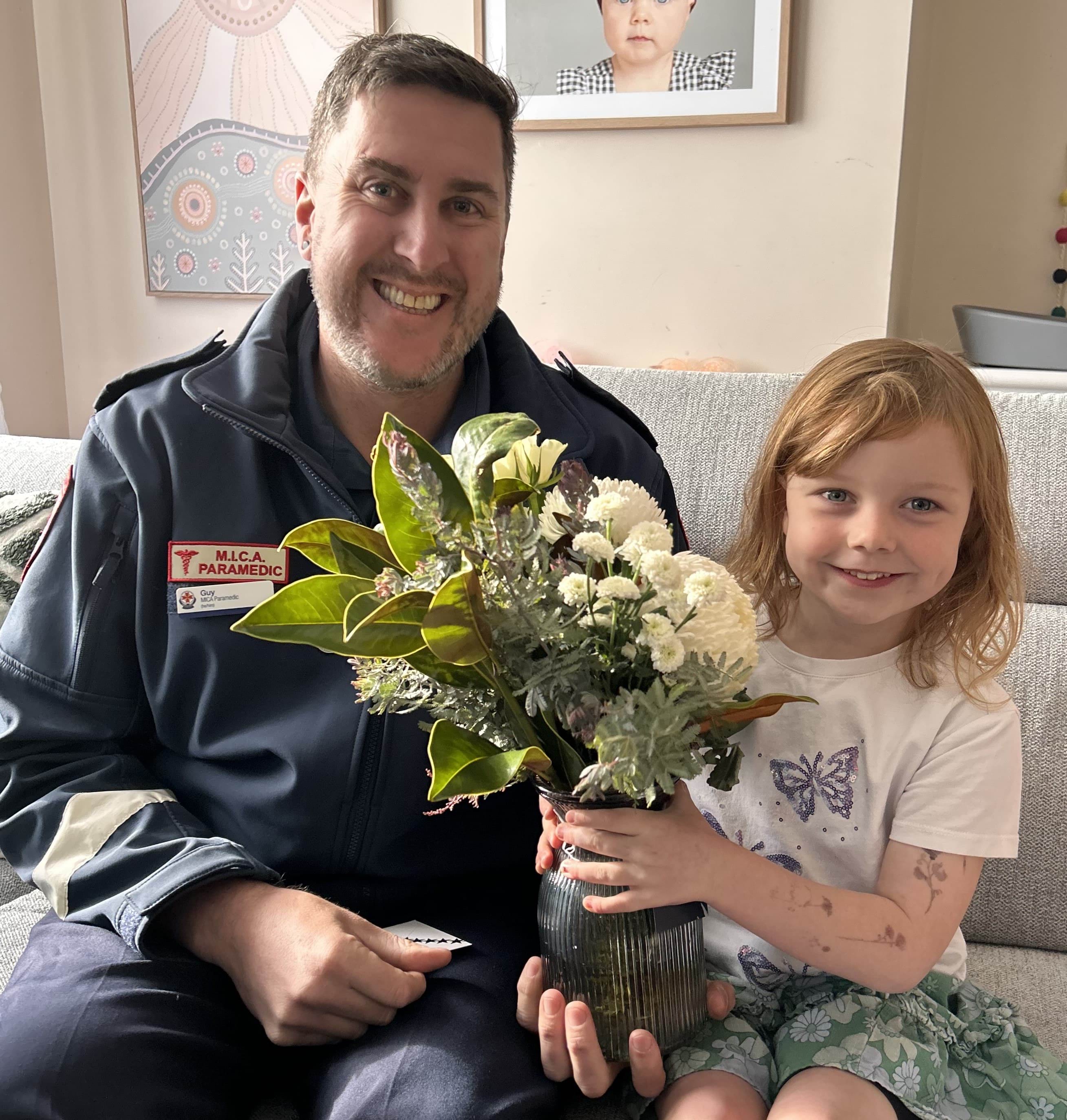 A smiling male paramedic receiving flowers from a smiling five year old girl.