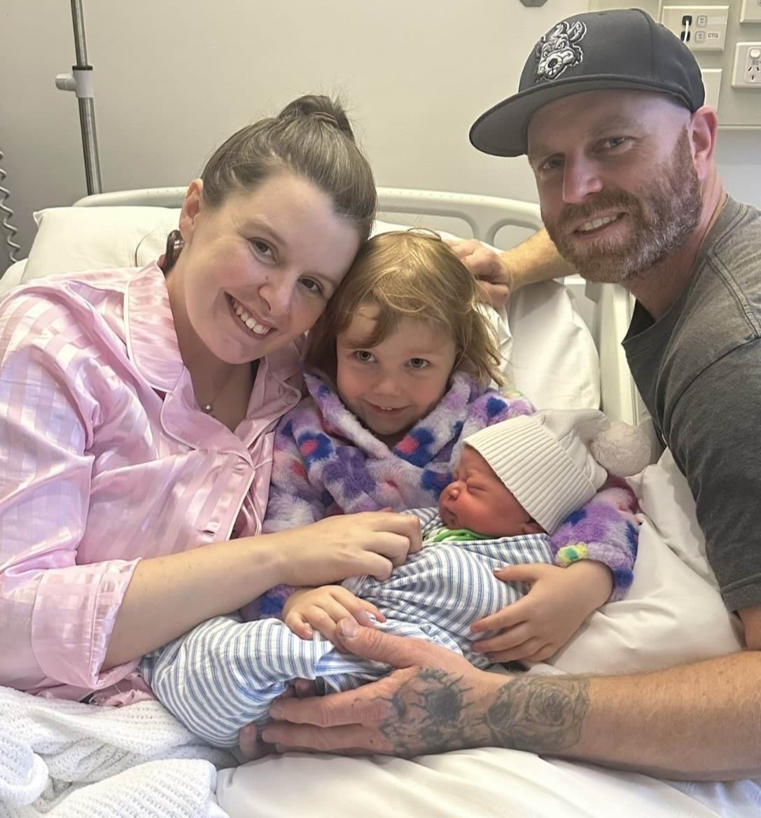 A woman, young girl holding a baby and man smiling at the camera from a hospital bed.