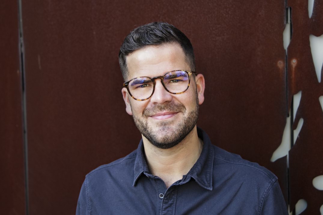 A man with brown hair and short facial hair smiles for the camera. He wears glasses.