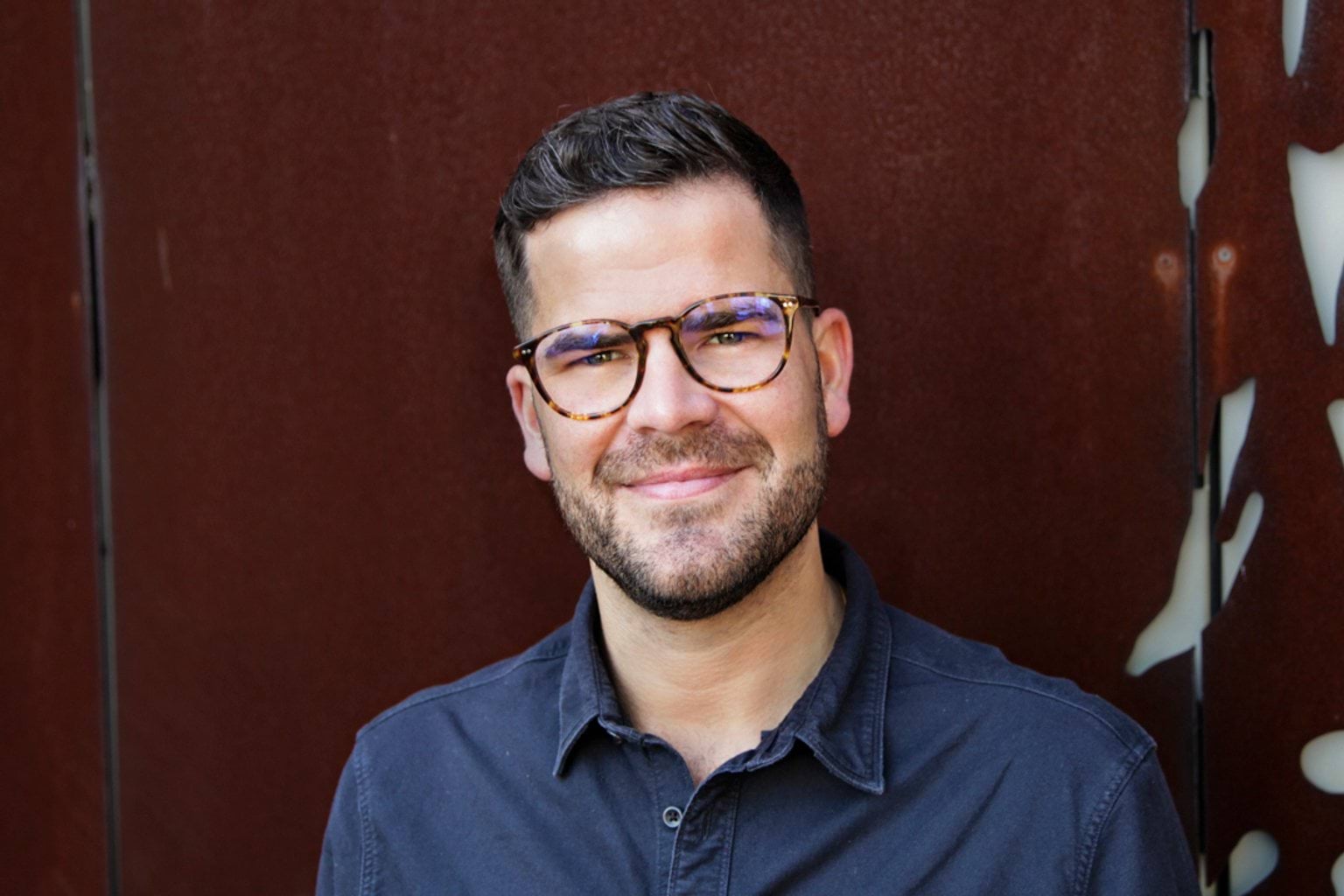A man with brown hair and short facial hair smiles for the camera. He wears glasses.