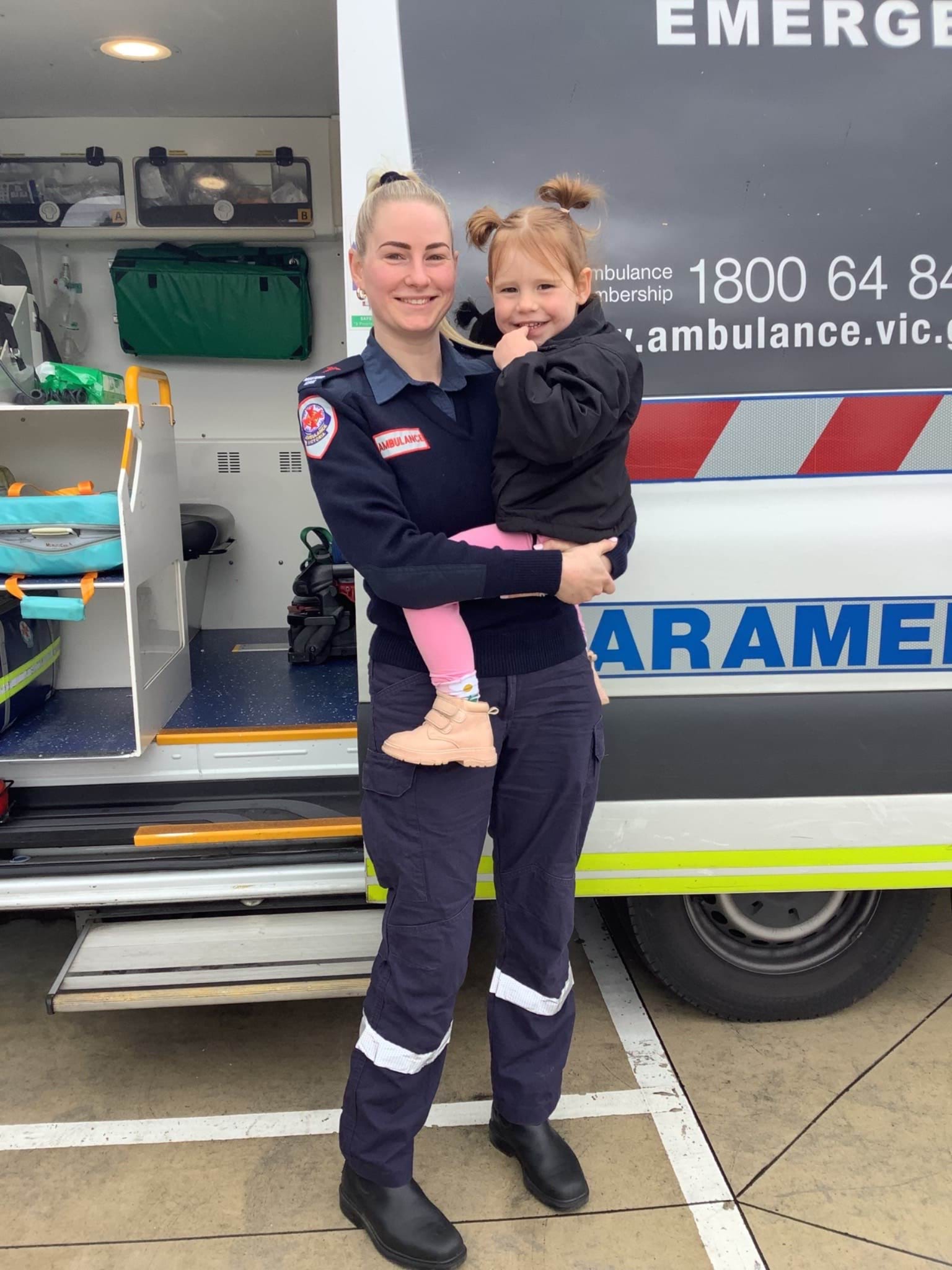 A woman in Ambulance Victoria uniform holds a little girl in front of an ambulance.