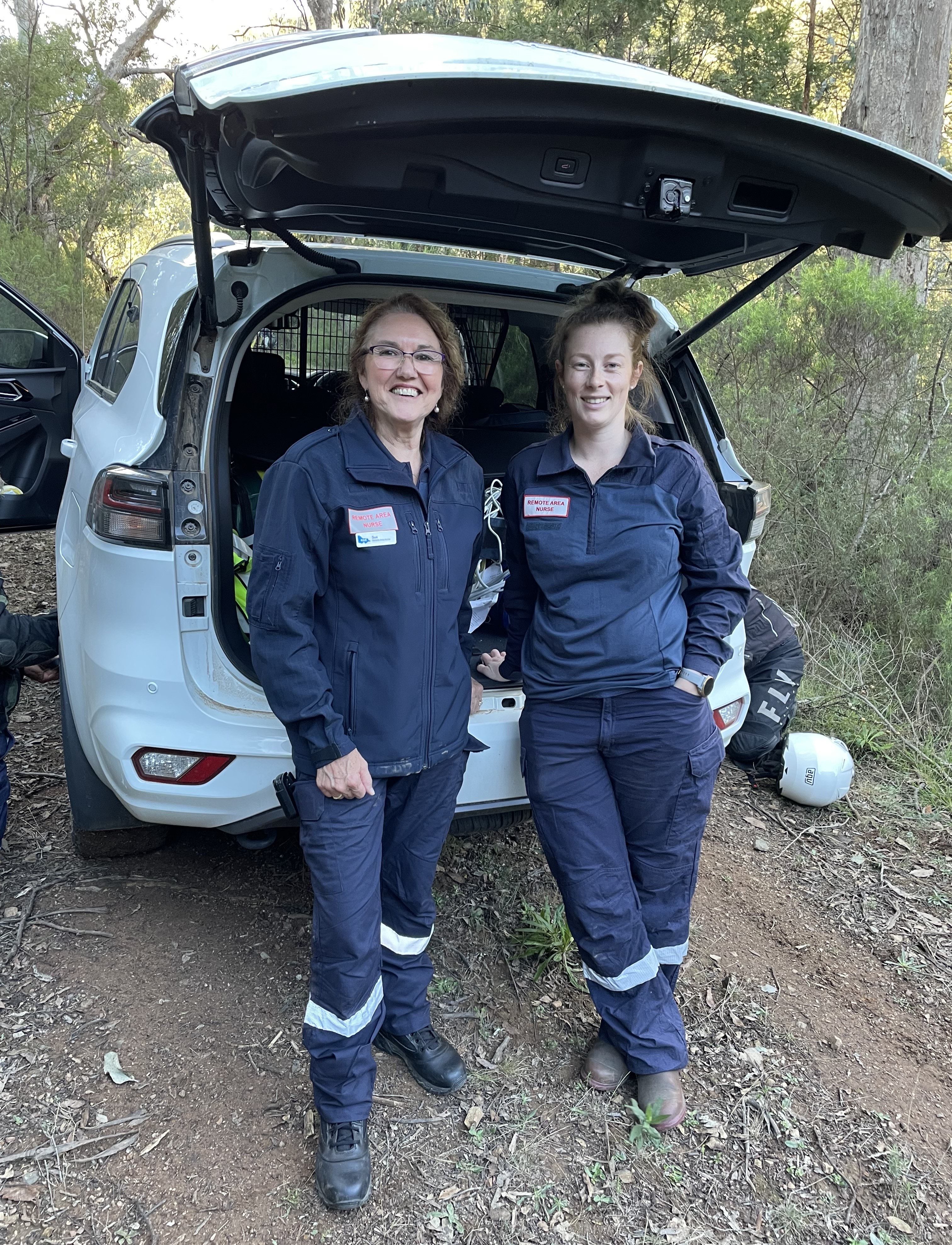 Two women in Remote Area Nurse uniform stand behind a car on a bush track.