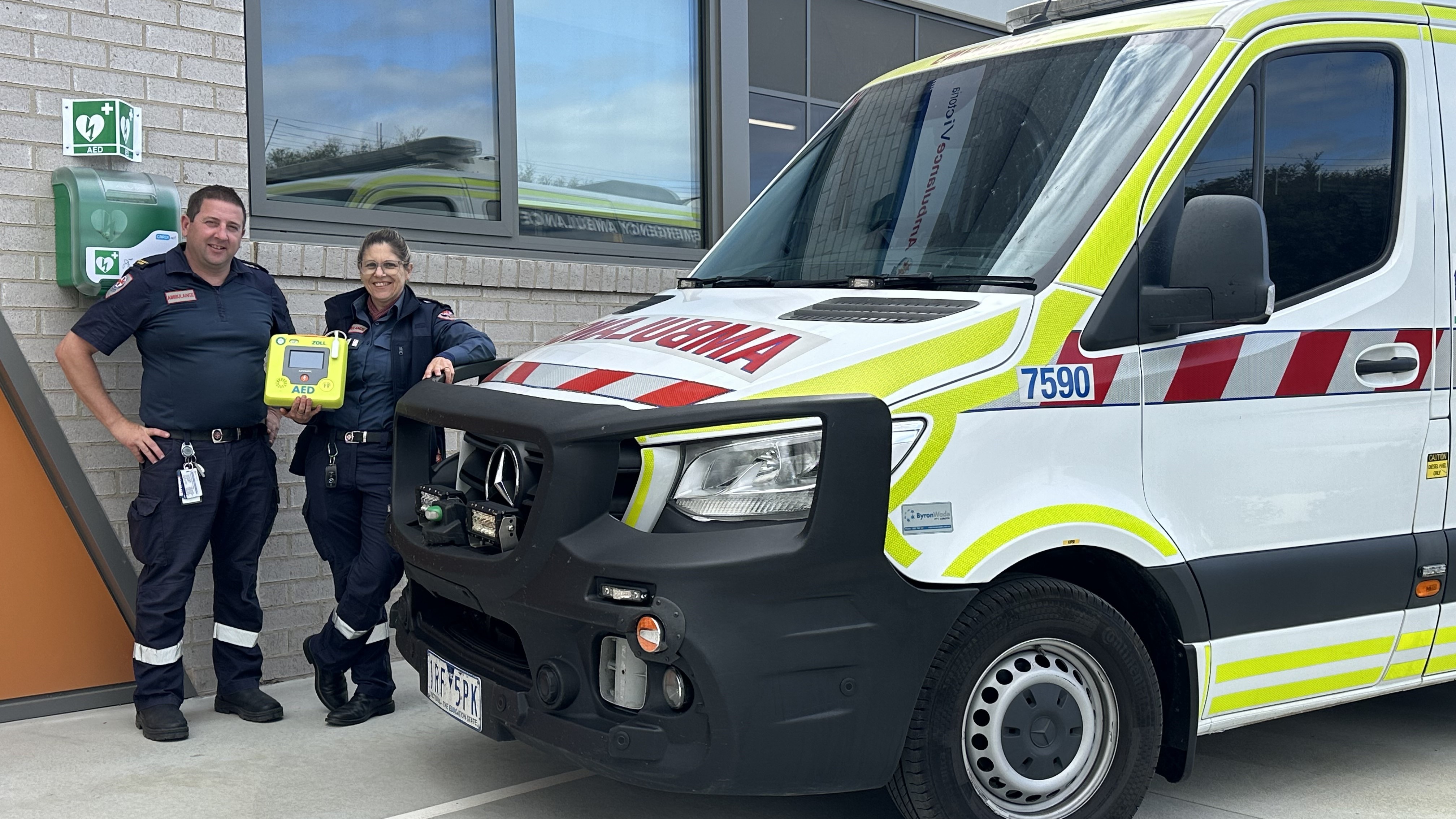 A man and woman holding an automated external defibrillator standing next to an ambulance.
