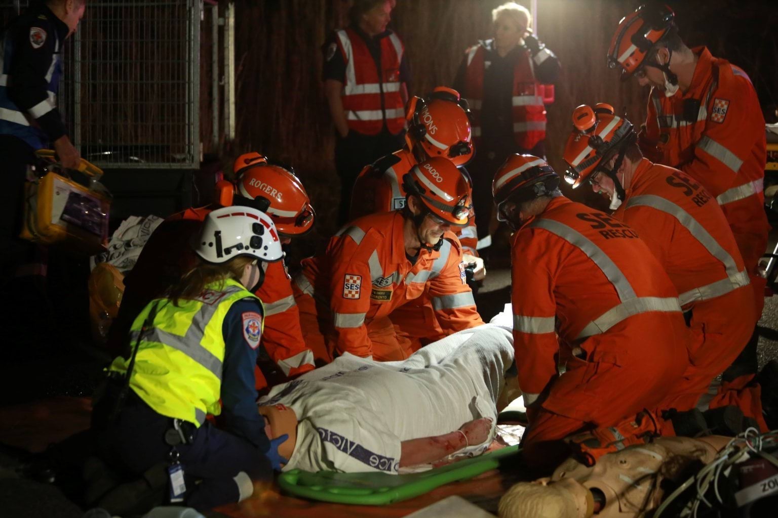 A paramedic in a yellow high-vis vest and SES volunteers in orange lift a patient on a stretcher.