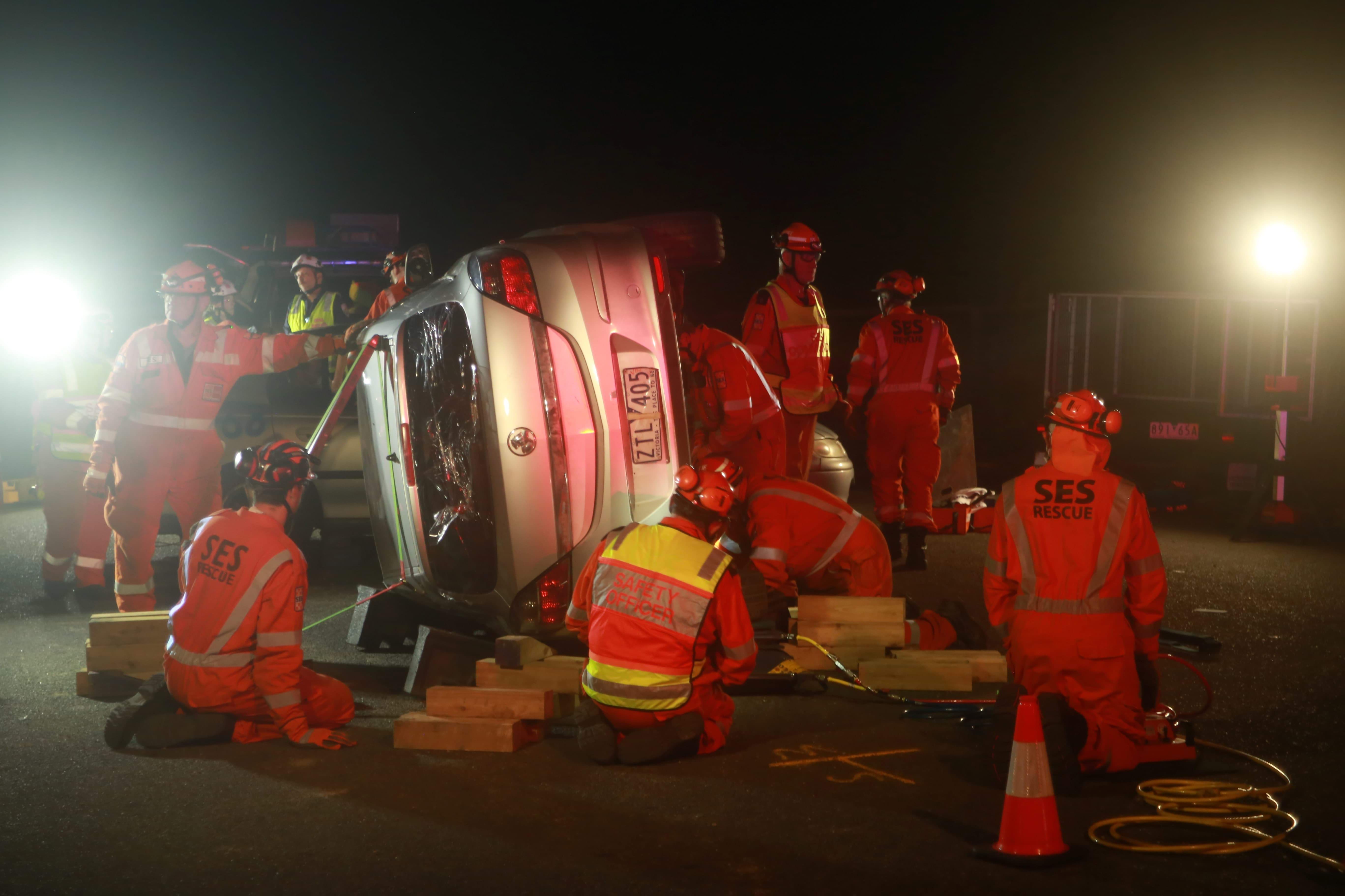 SES volunteers in orange surround a silver car that rests on its side. The back window of the car is shattered.