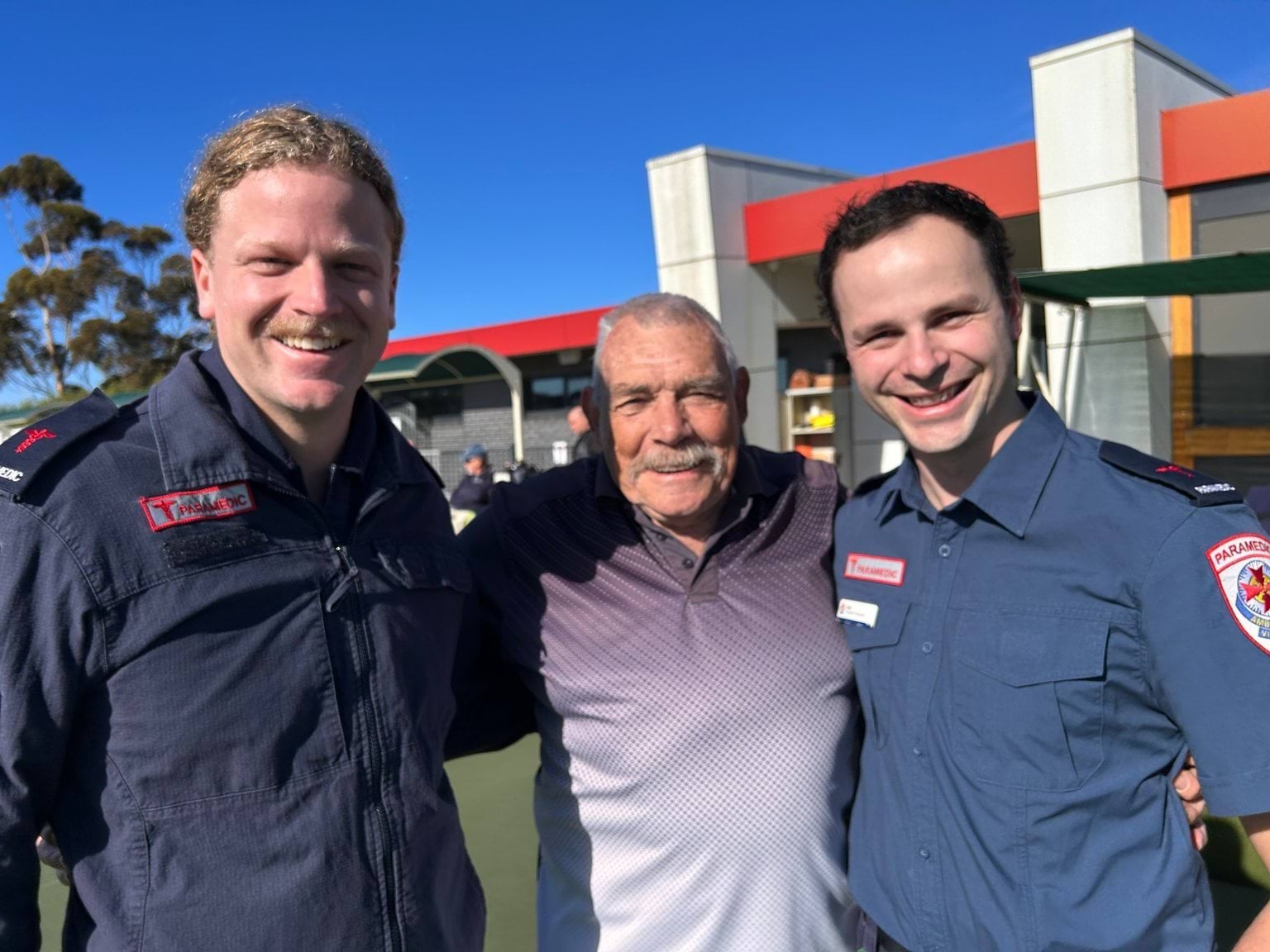 two smiling paramedics standing either side of a smiling 80 year old man. 