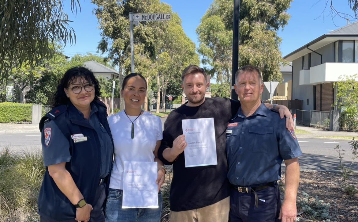 Two paramedics stand with their arms around two people in casual clothes holding certificates.