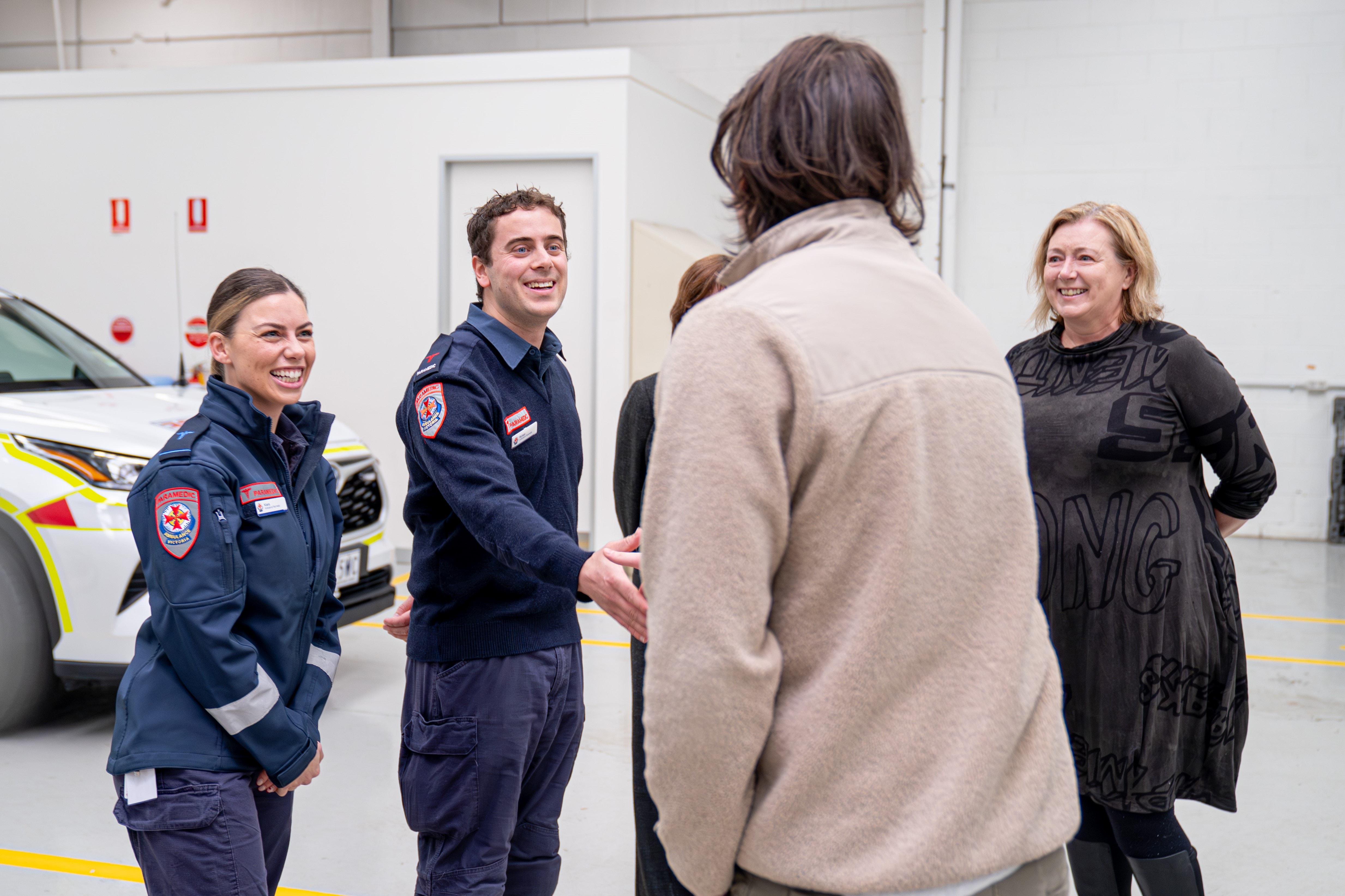 Two paramedics smile and greet a man with his back to camera. A woman stands next to them and smiles at the group.