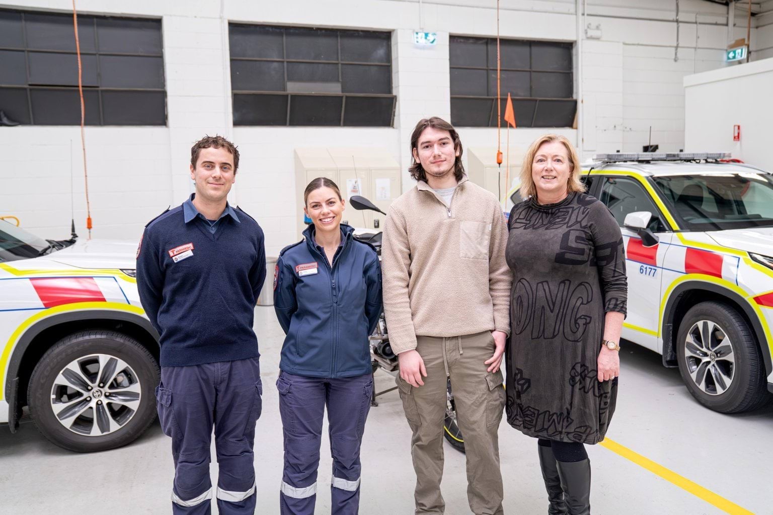 Two paramedics stand next to a man and woman in casual clothes. Ambulance vehicles are behind them.