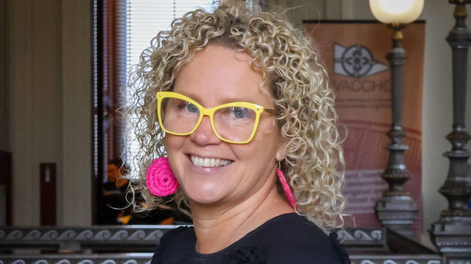 A women with bright yellow glasses and pink woven earrings poses for a corporate portrait