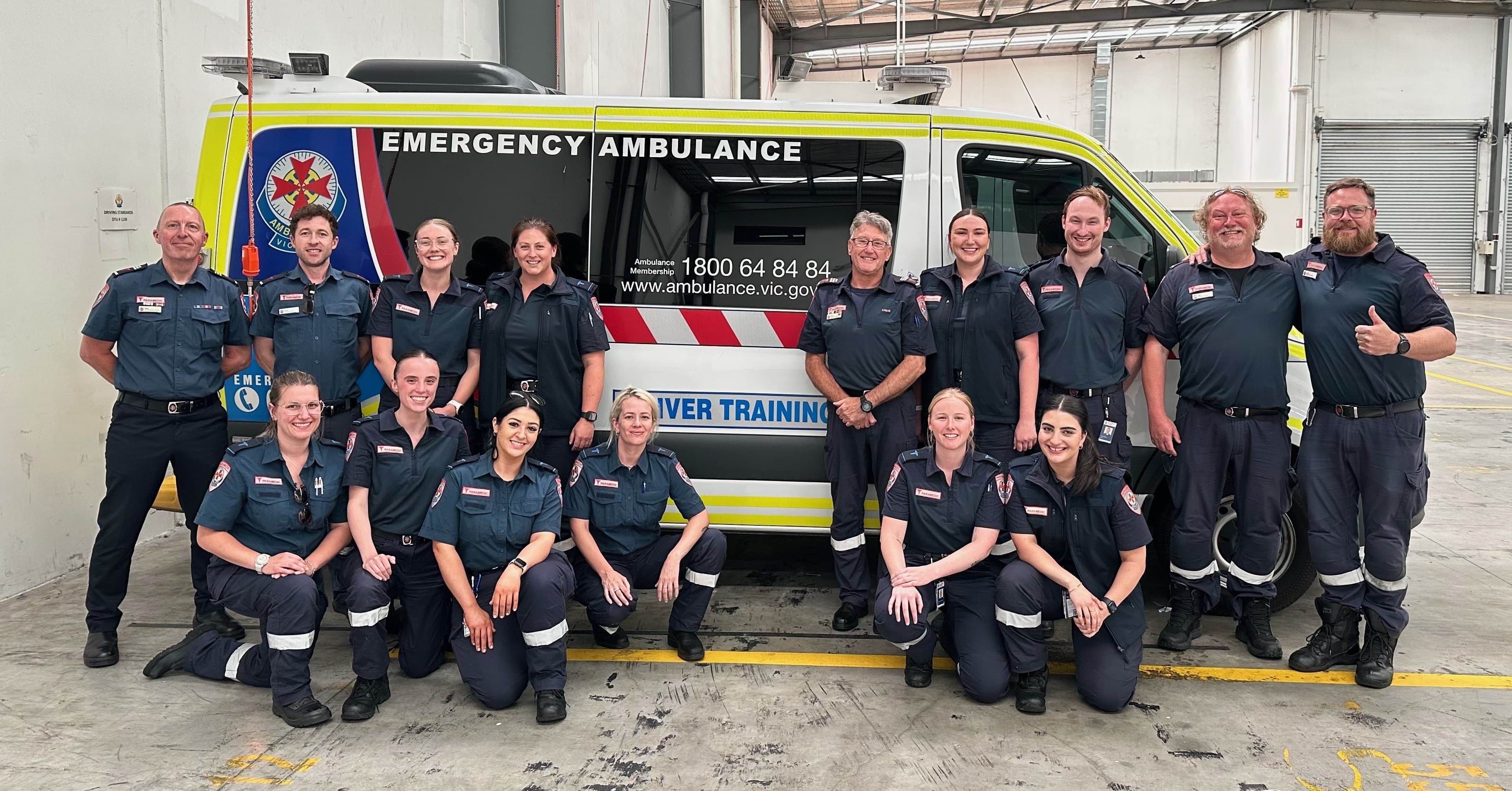 A group of 15 people in Ambulance Victoria uniform smile for the camera in front of a 'Driver Training' ambulance.