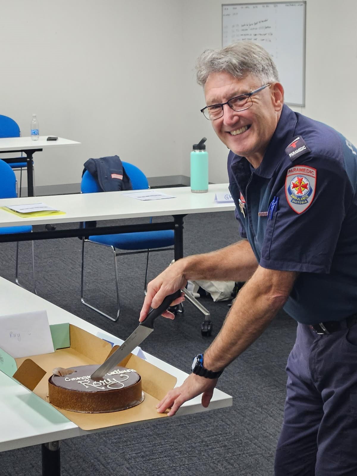 A man in Ambulance Victoria paramedic uniform smiles for the camera while cutting a cake.