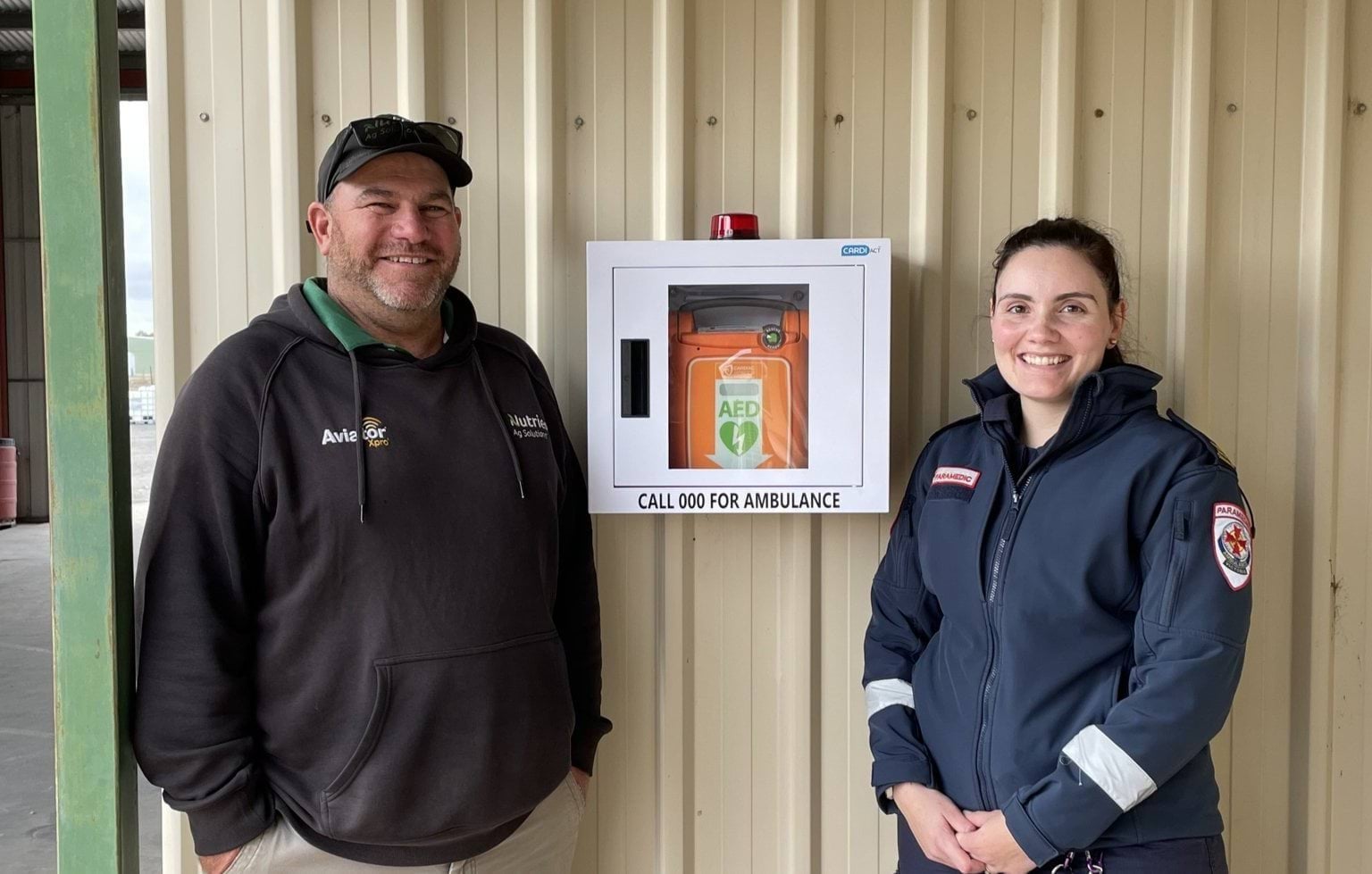A man and a woman smiling while standing either side of a wall mounted automated external defibrillator (AED).