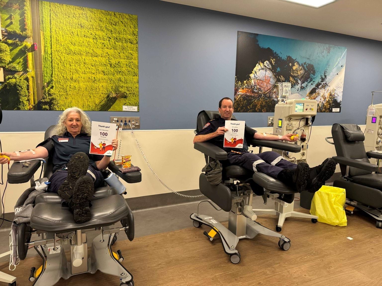 A woman and a man sitting down donating plasma holding up 100 donation certificates.
