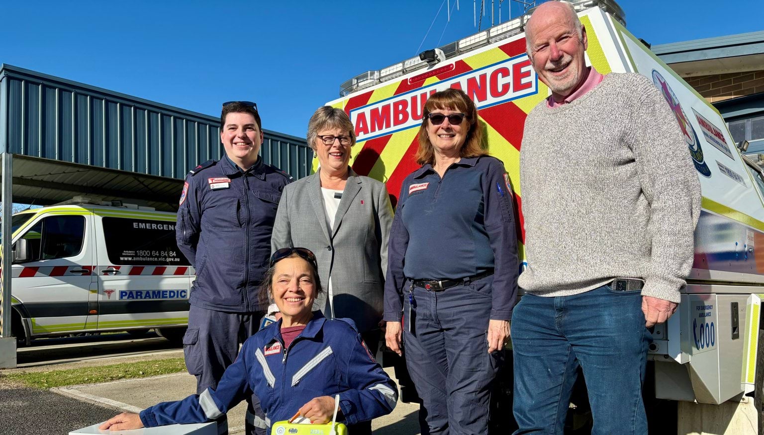 Three people in Ambulance Victoria uniform and two people in casual clothes smile for the camera in front of an ambulance.