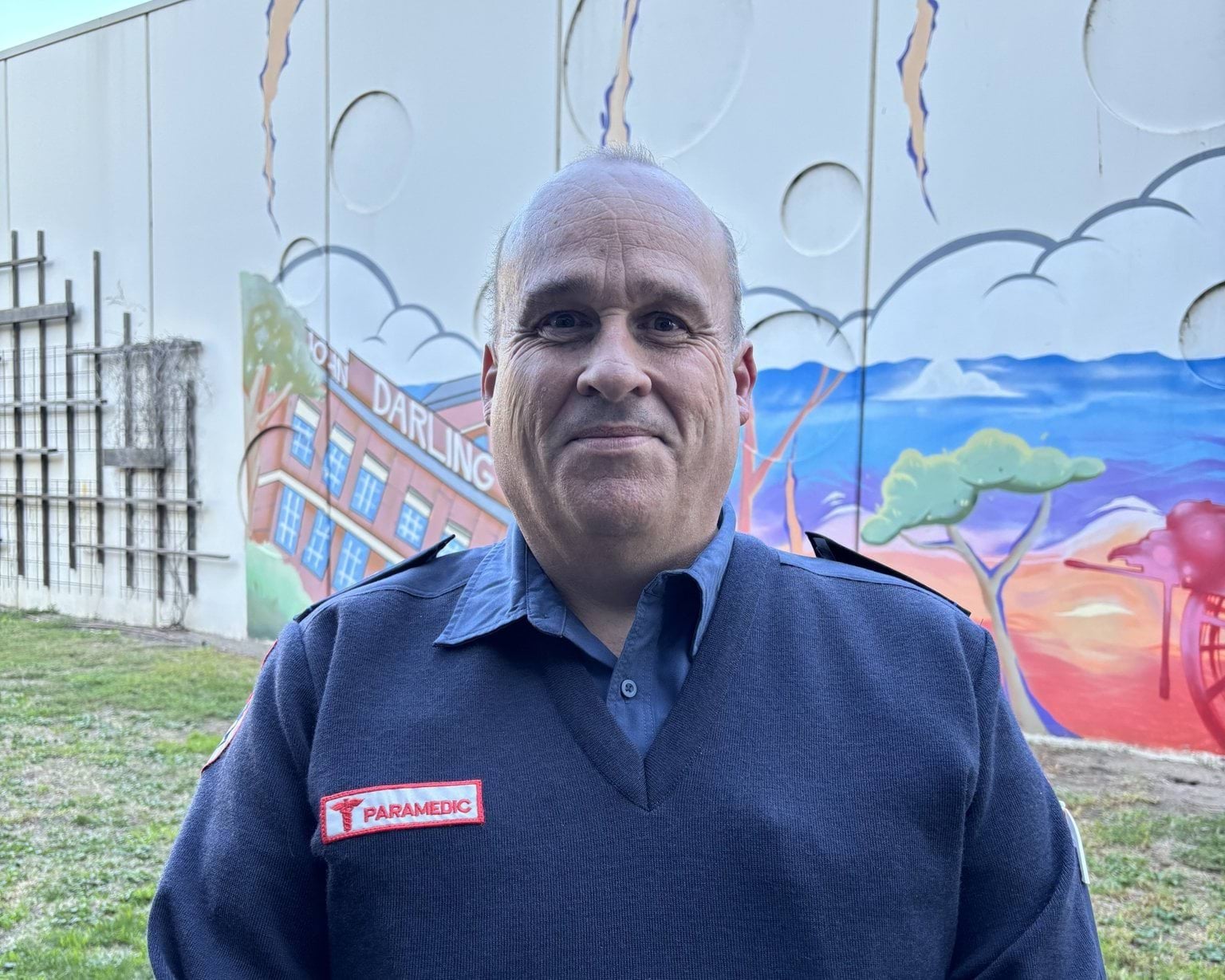 A headshot of a man in paramedic uniform in front of a colourful mural on a wall.