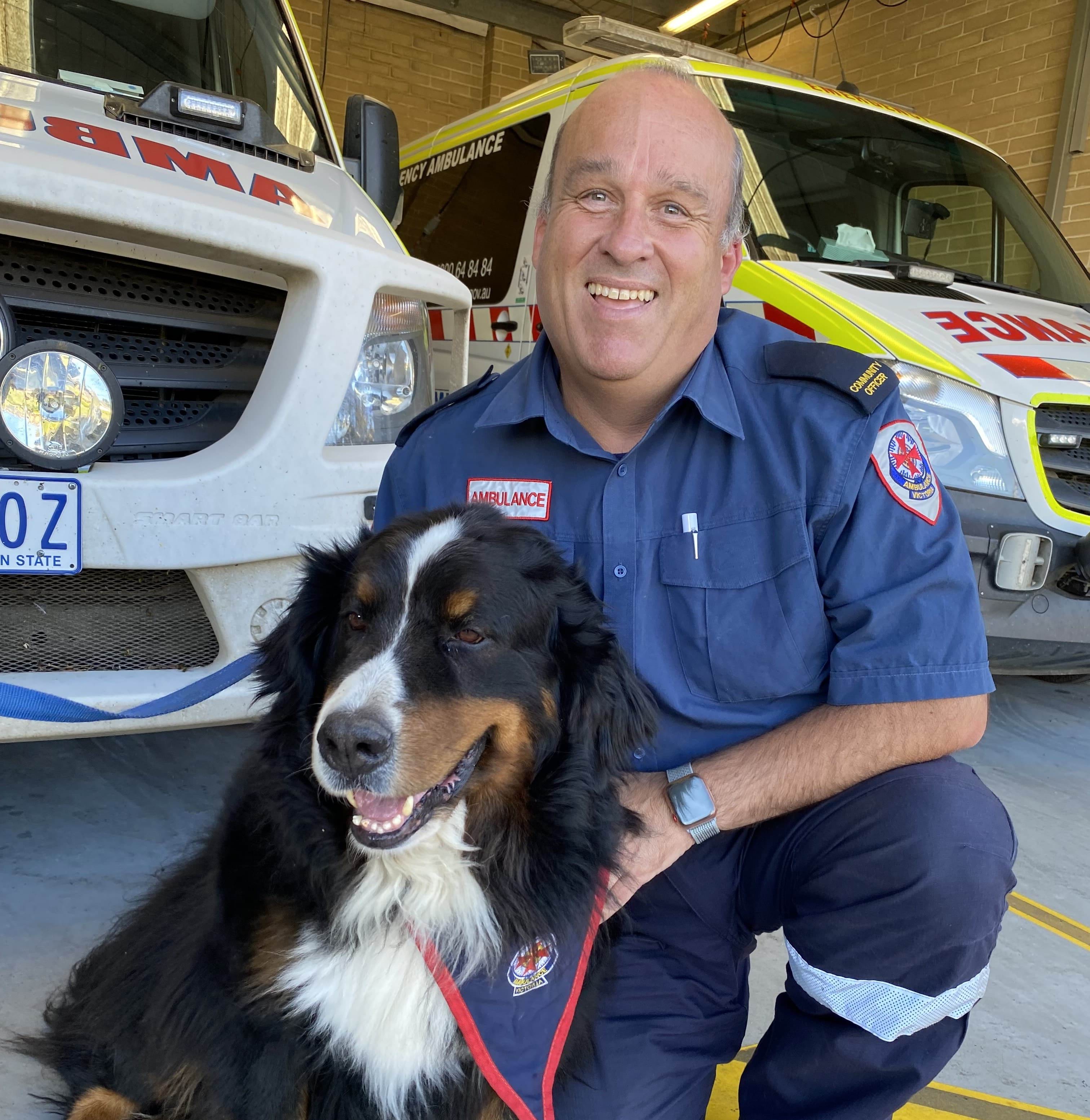 A man in Ambulance Victoria Ambulance Community Officer uniform smiles for the camera while patting a dog. Ambulance vehicles are in the background.