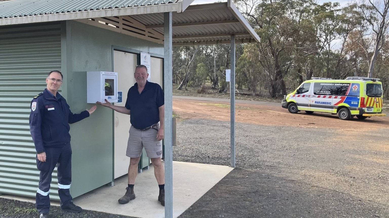 Two men pointing at an AED mounted on the wall of a toilet block.
