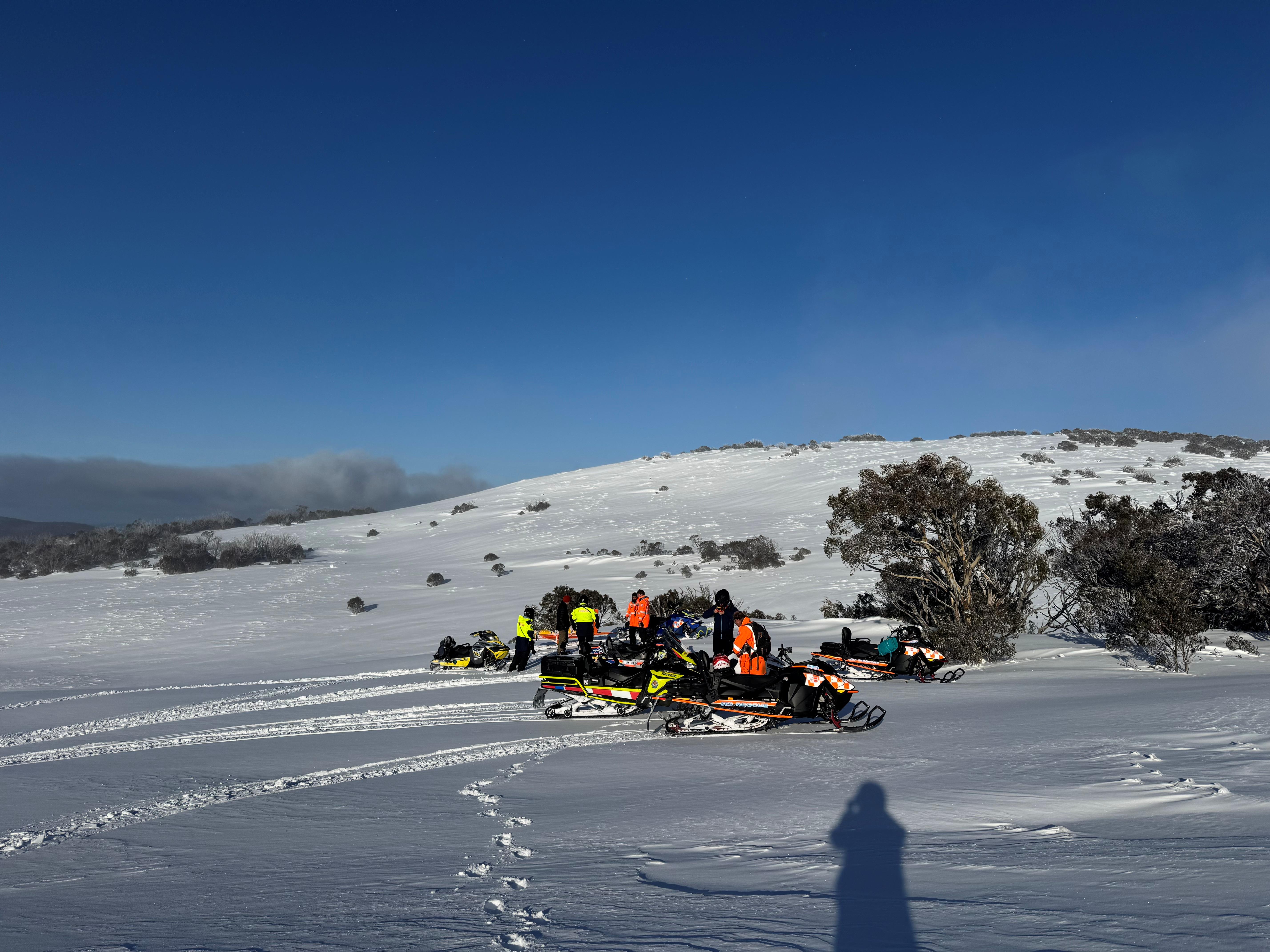 A group of emergency services personnel and their Skidoos in the snow. There is clear blue sky.