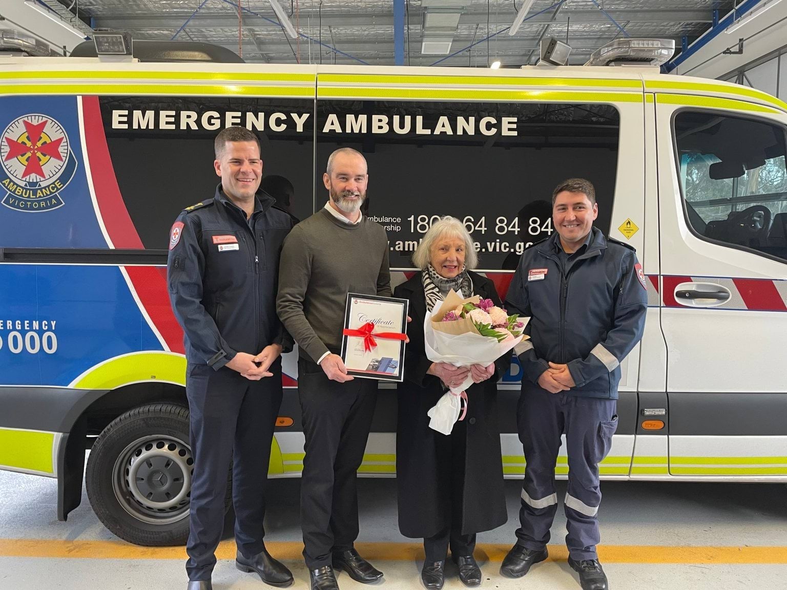 A man and woman hold a certificate and flowers. They stand between two men in paramedic uniform, in front of an Ambulance Victoria ambulance vehicle.