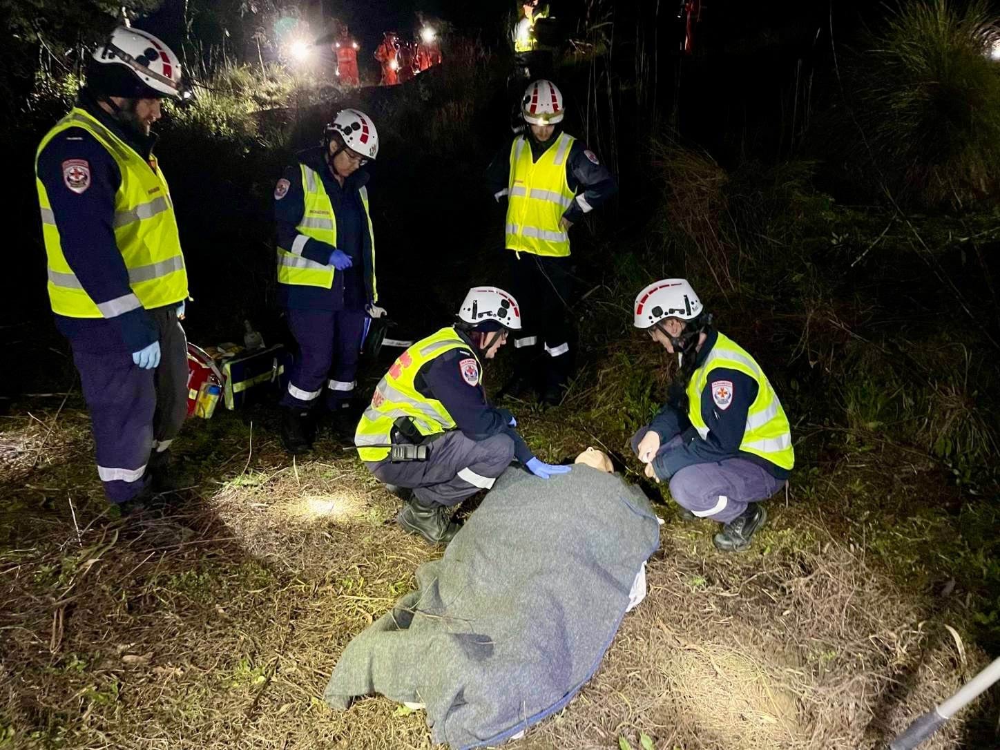 Five paramedics in high vis vests gather around a mannequin on the ground.