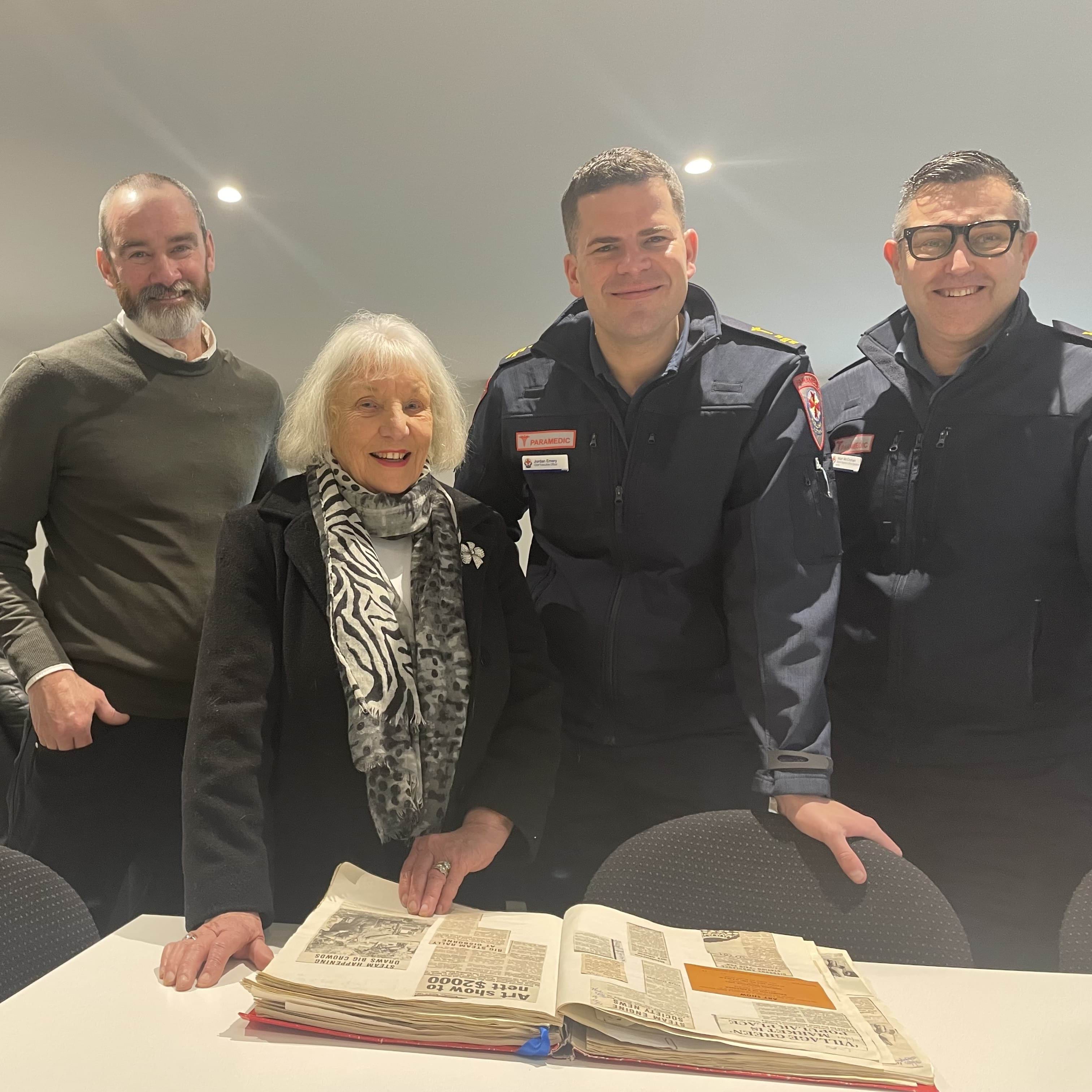 A woman is about to flip the page of a scrapbook with old newspaper clippings. She smiles for the camera with a man in casual clothes and two paramedics.