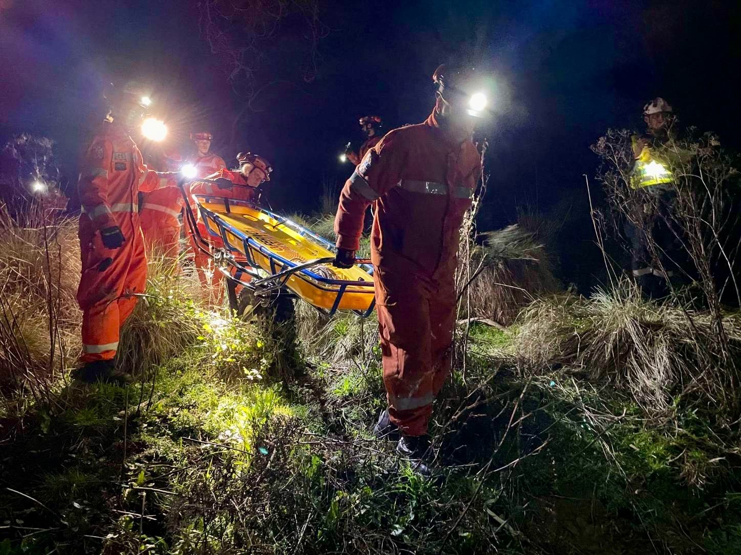 SES volunteers wheel a stretcher down a grassy, bushy hill at night.